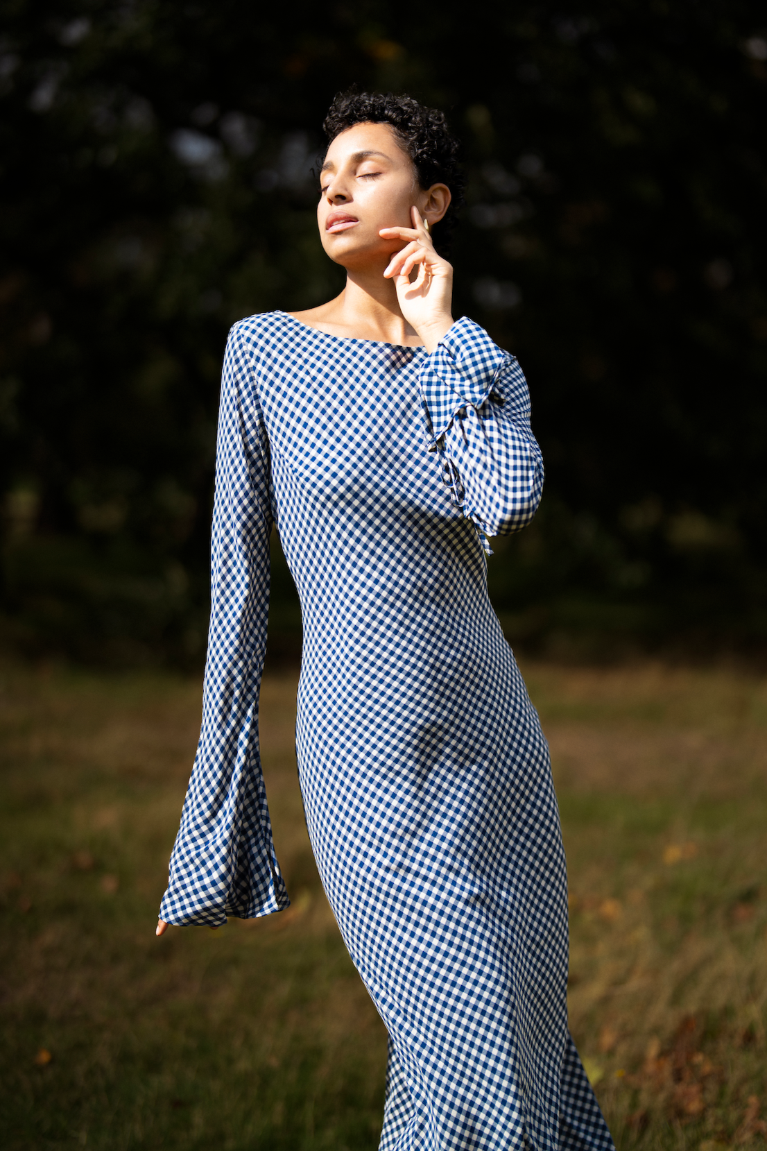 Woman in a blue checkered dress standing in a natural setting with trees and grass.
