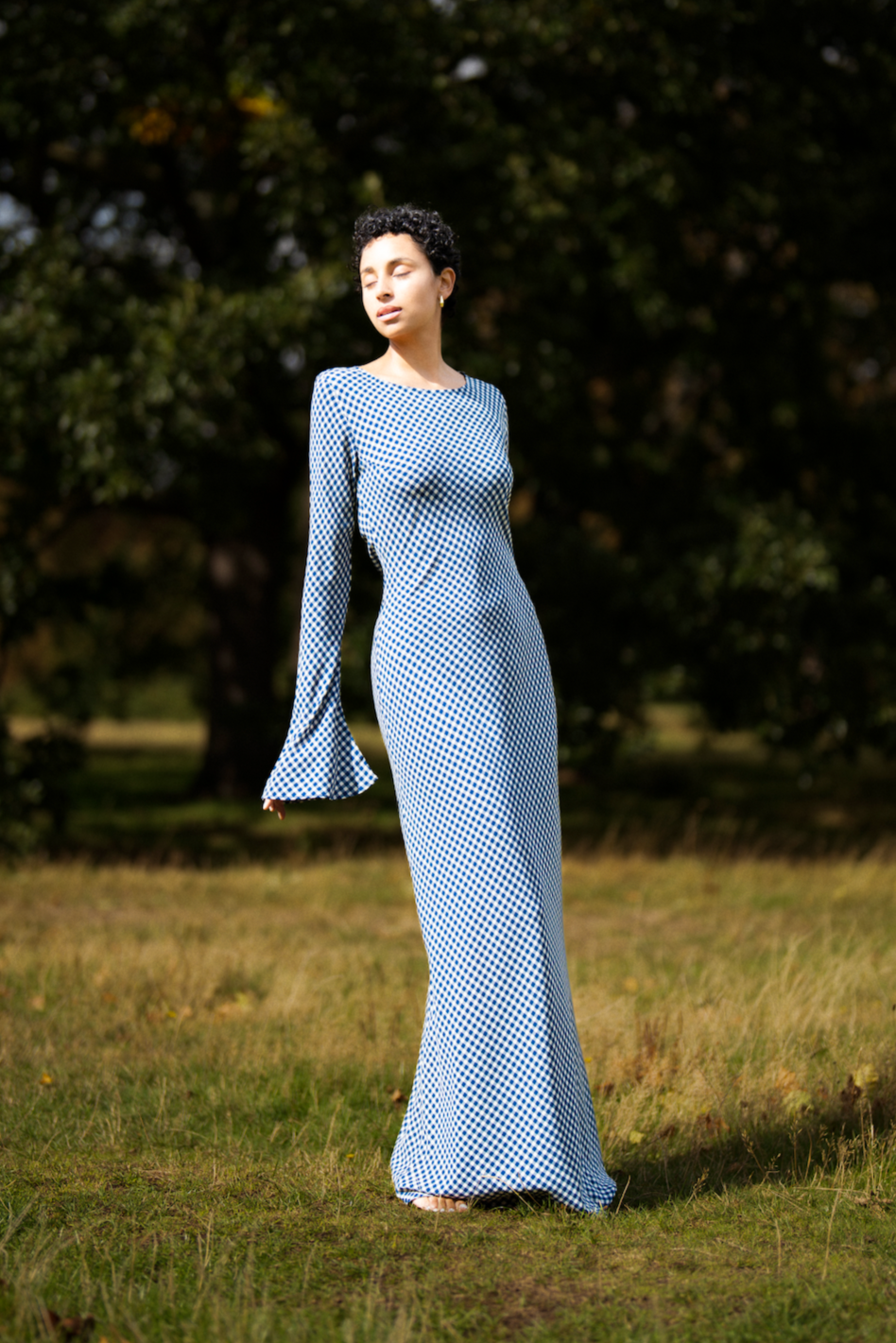 Woman in a long, light blue dress standing in a grassy field with trees in the background